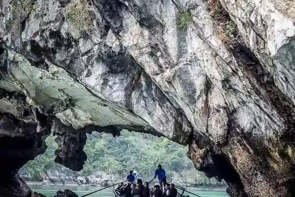 Wooden boat with tourists rowing through limestone arch over emerald sea.