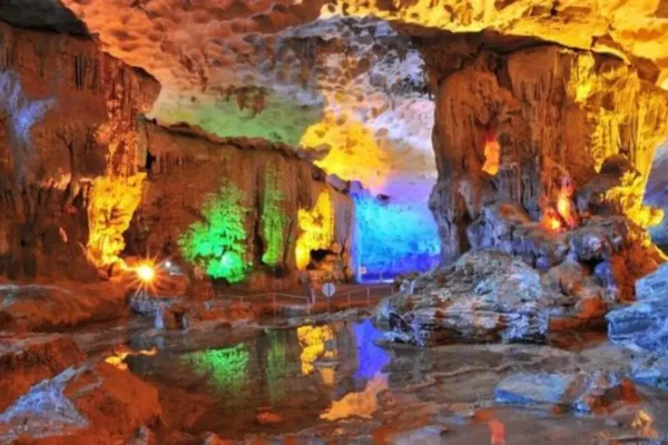 Colorfully lit cave interior with stalactites, stalagmites, and a reflective water surface