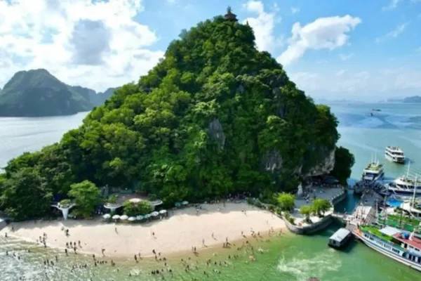 Tourists swimming and relaxing on a sandy beach at the base of a green mountain on Ti Top Island, with boats docked nearby